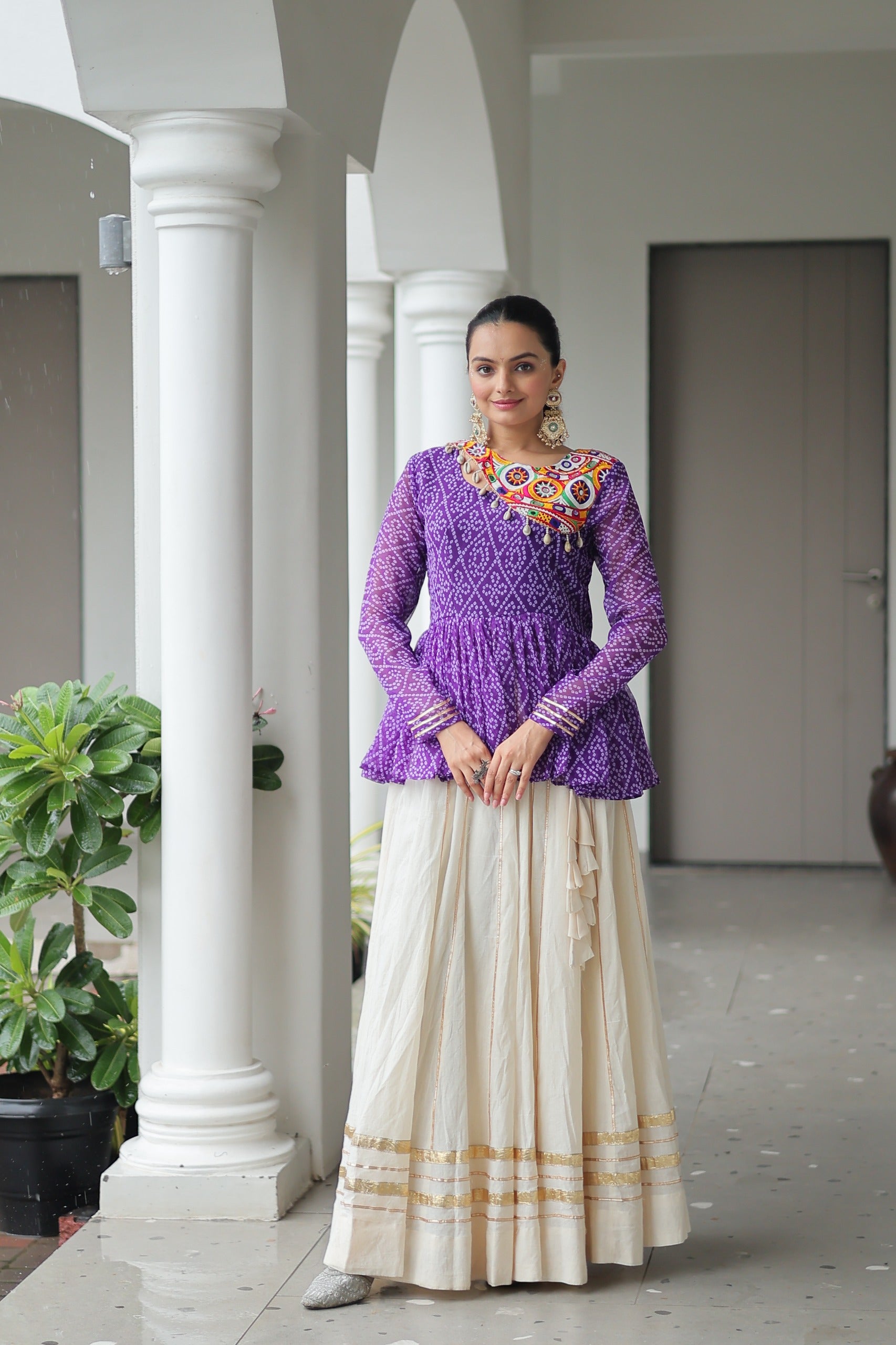 Woman in a purple top and white skirt standing in a colonnaded area with plants.