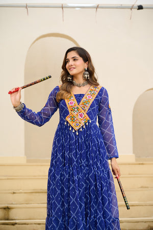 Woman in a blue traditional outfit with intricate designs, holding a decorative stick against a beige wall.