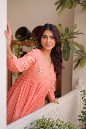 Woman in a peach-colored dress with floral patterns standing indoors with plants around.