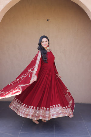 Woman in a red traditional outfit with white embroidery standing against a beige wall.