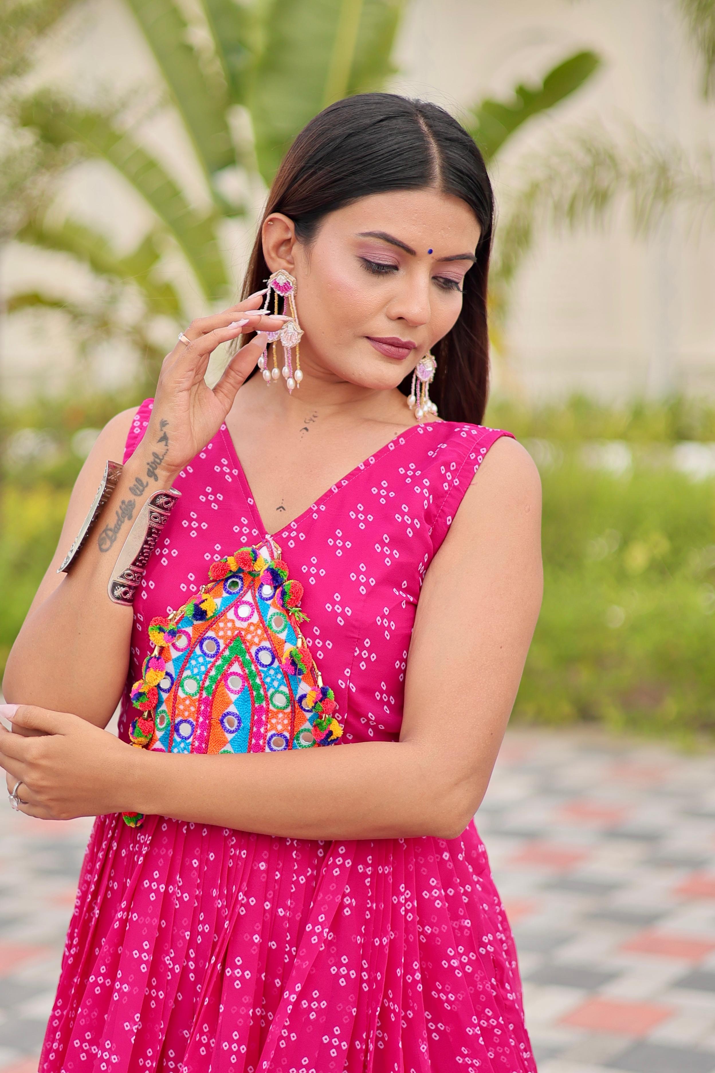 Woman in a pink traditional outfit with a colorful blouse, standing outdoors with greenery in the background.