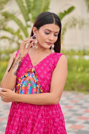 Woman in a pink traditional outfit with a colorful blouse, standing outdoors with greenery in the background.