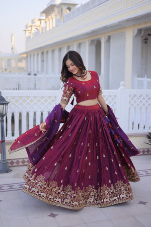 Woman in a traditional maroon and purple embroidered outfit standing in front of a white building.