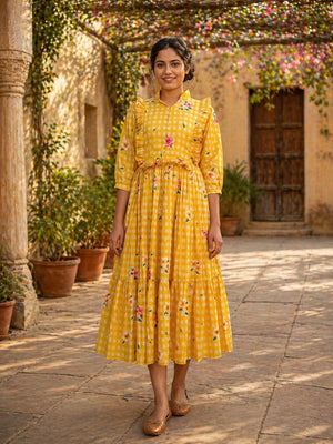 Woman in a yellow checkered dress standing in an outdoor setting with plants and a building in the background.