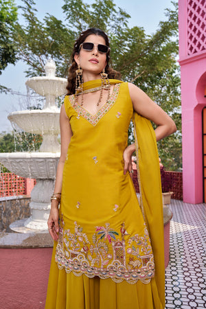 Woman in a yellow traditional outfit with intricate designs, standing outdoors with a fountain and pink building in the background.
