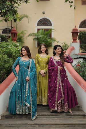 Three women in traditional dresses standing on steps with a building and plants in the background