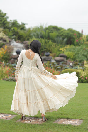 Woman in a long, flowing white dress standing in a garden with greenery and a pond in the background.