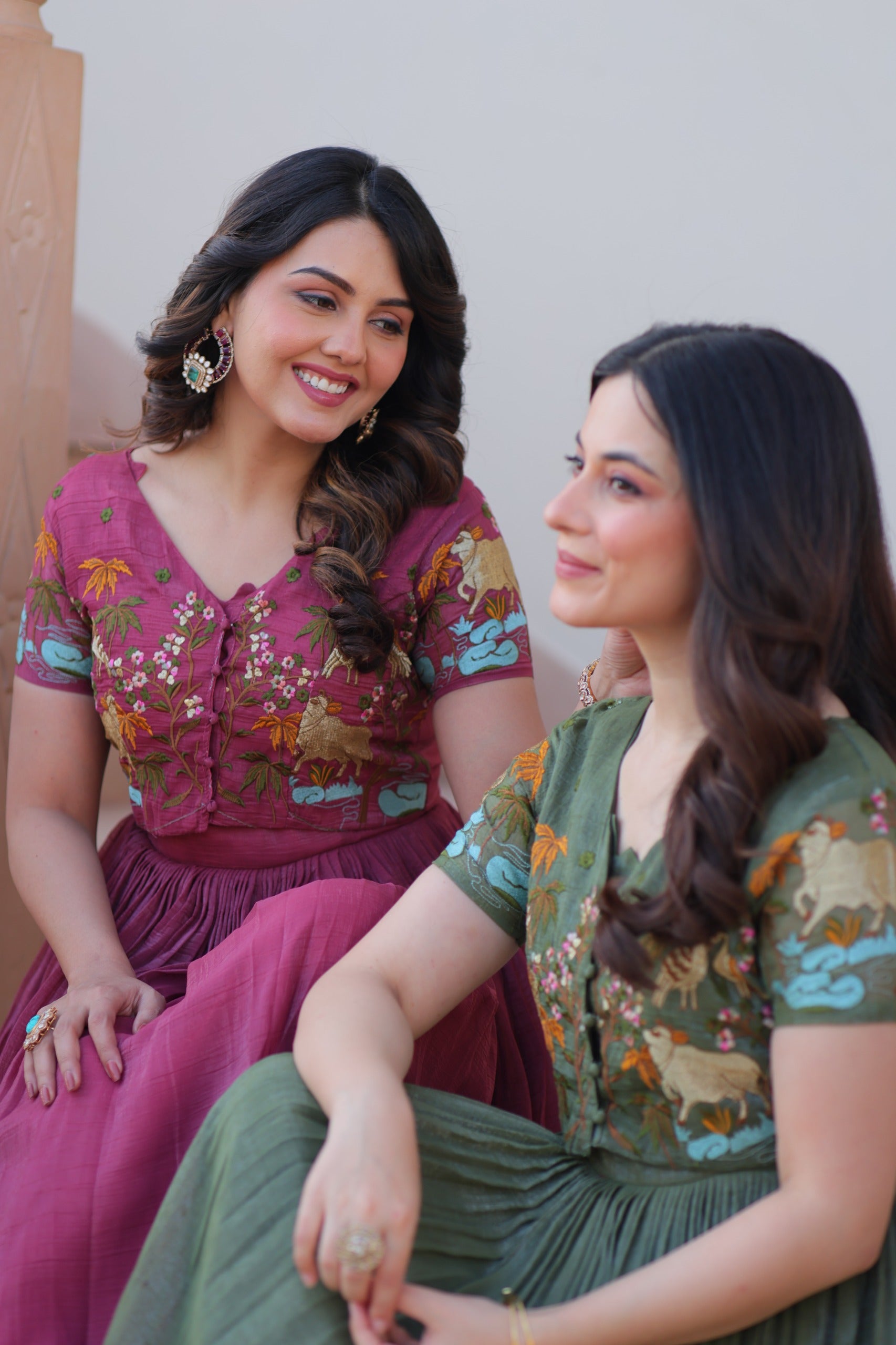 Two women in traditional outfits sitting and smiling against a neutral background