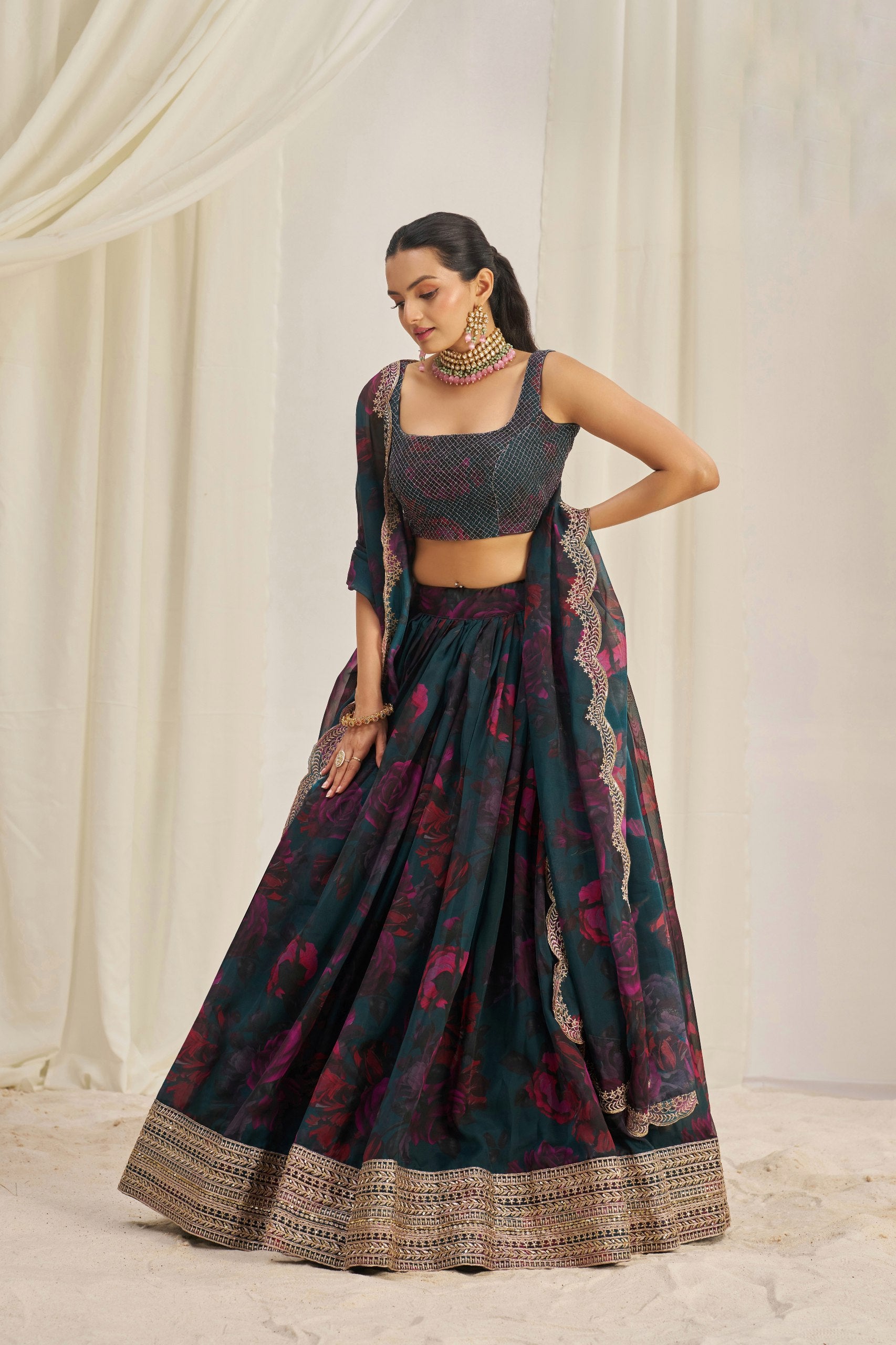 Woman wearing a traditional dark green and red lehenga with intricate patterns against a white curtain background.