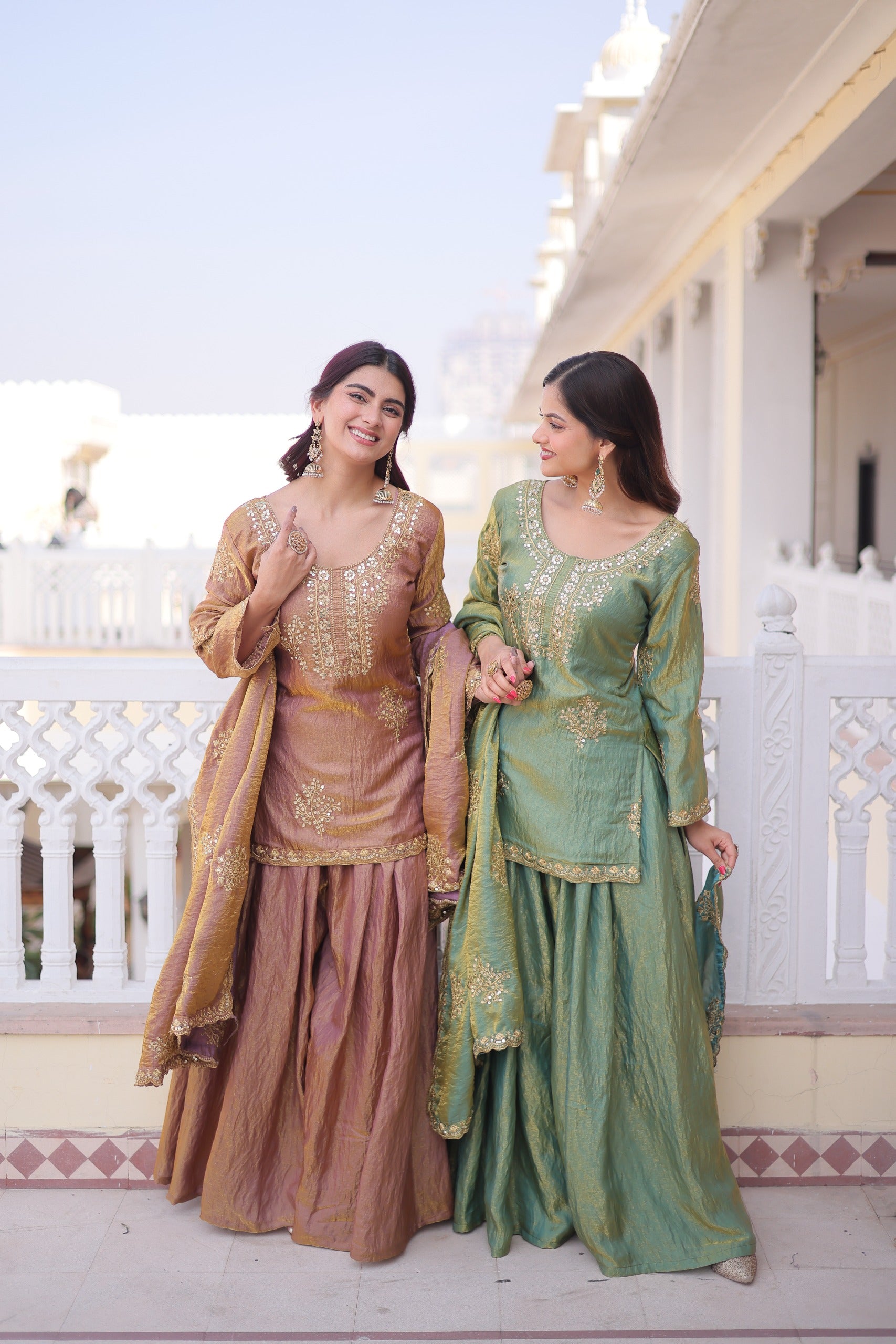 Two women in traditional outfits standing on a balcony with a white building in the background.