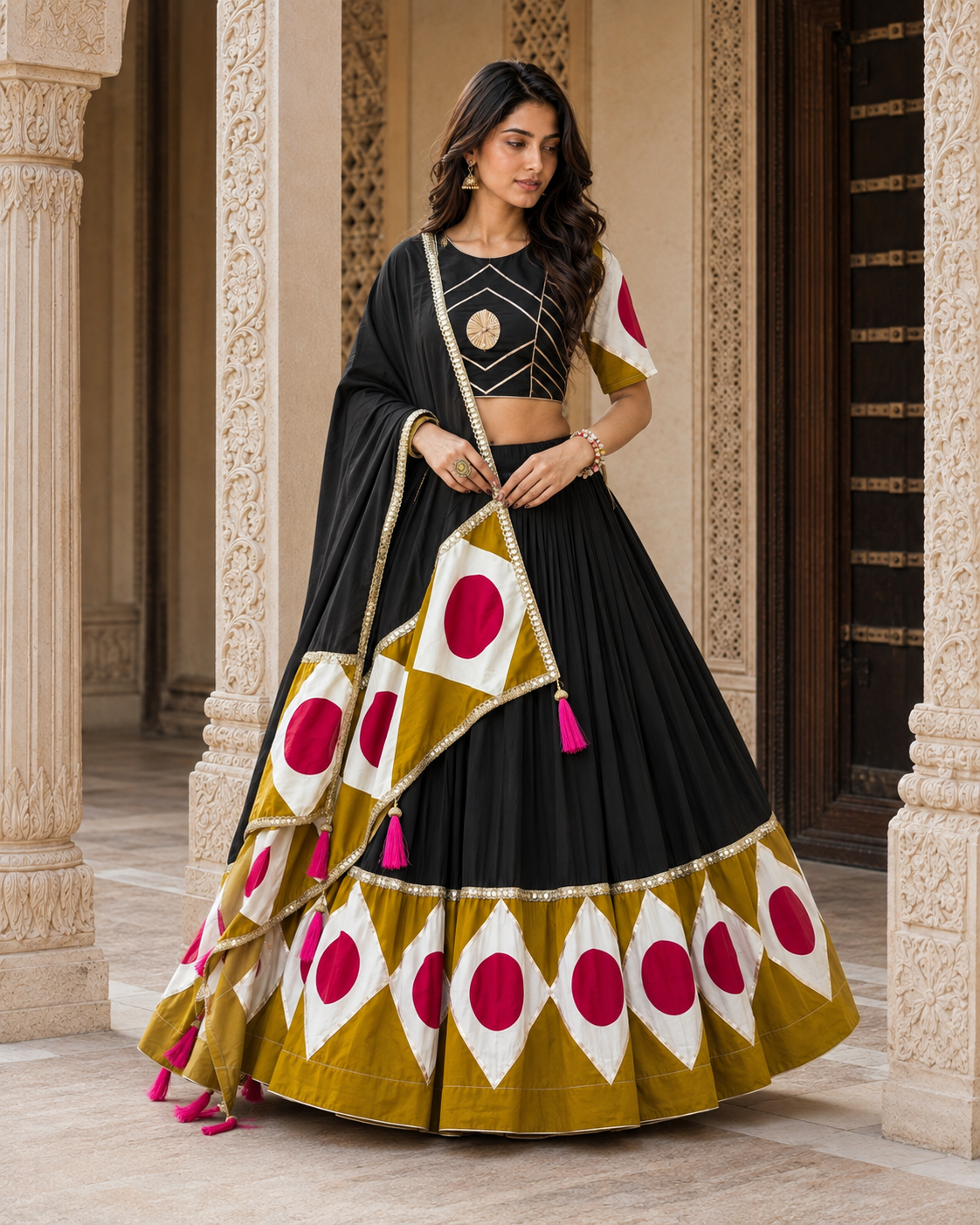 A woman models a traditional Indian lehenga choli set in an ornate architectural corridor. The outfit features a black skirt and blouse with gold embroidery, paired with a mustard yellow dupatta and flared hem decorated with white diamond panels containing magenta-pink circular motifs.