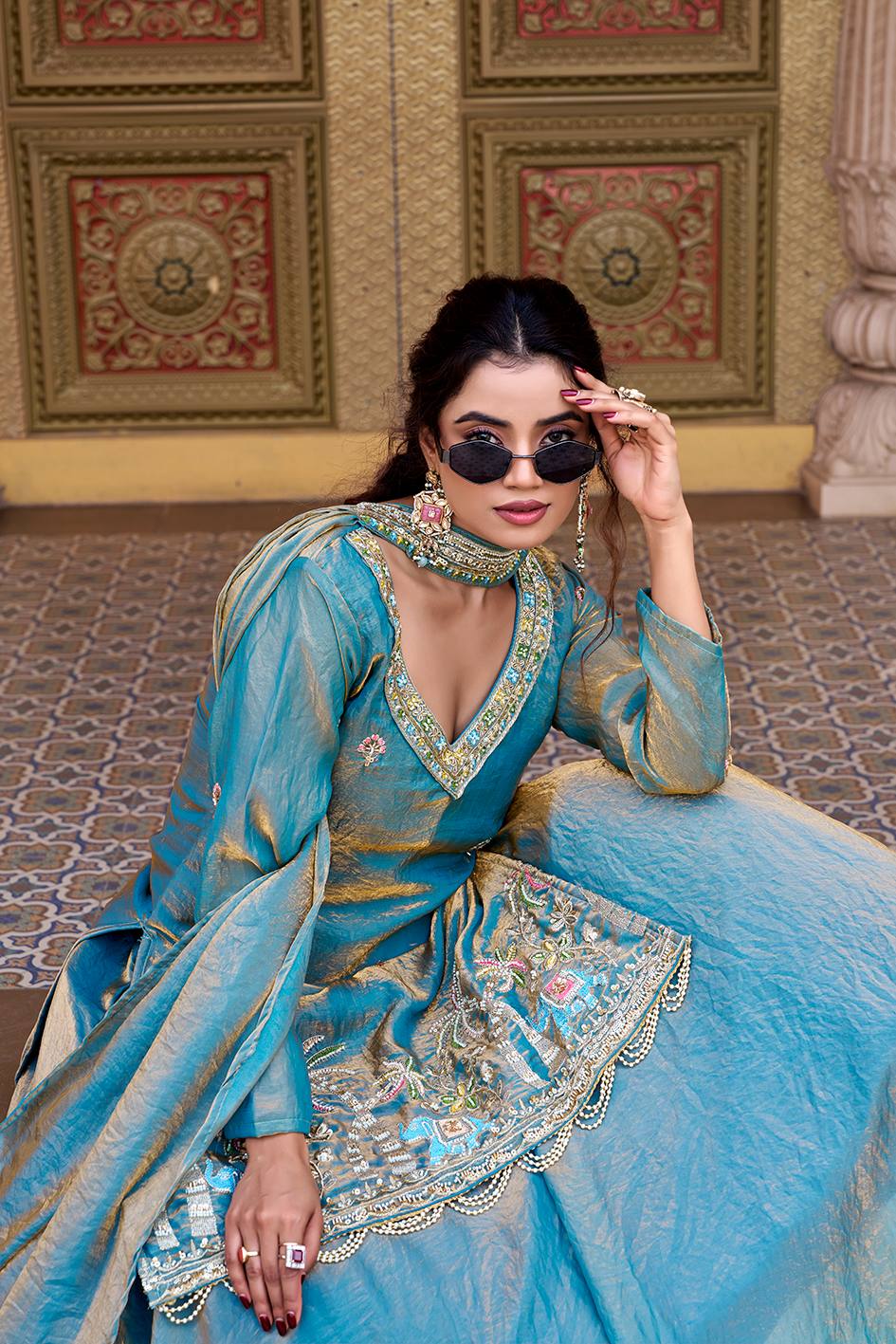 Woman in a blue traditional outfit with intricate designs, sitting on a patterned floor.