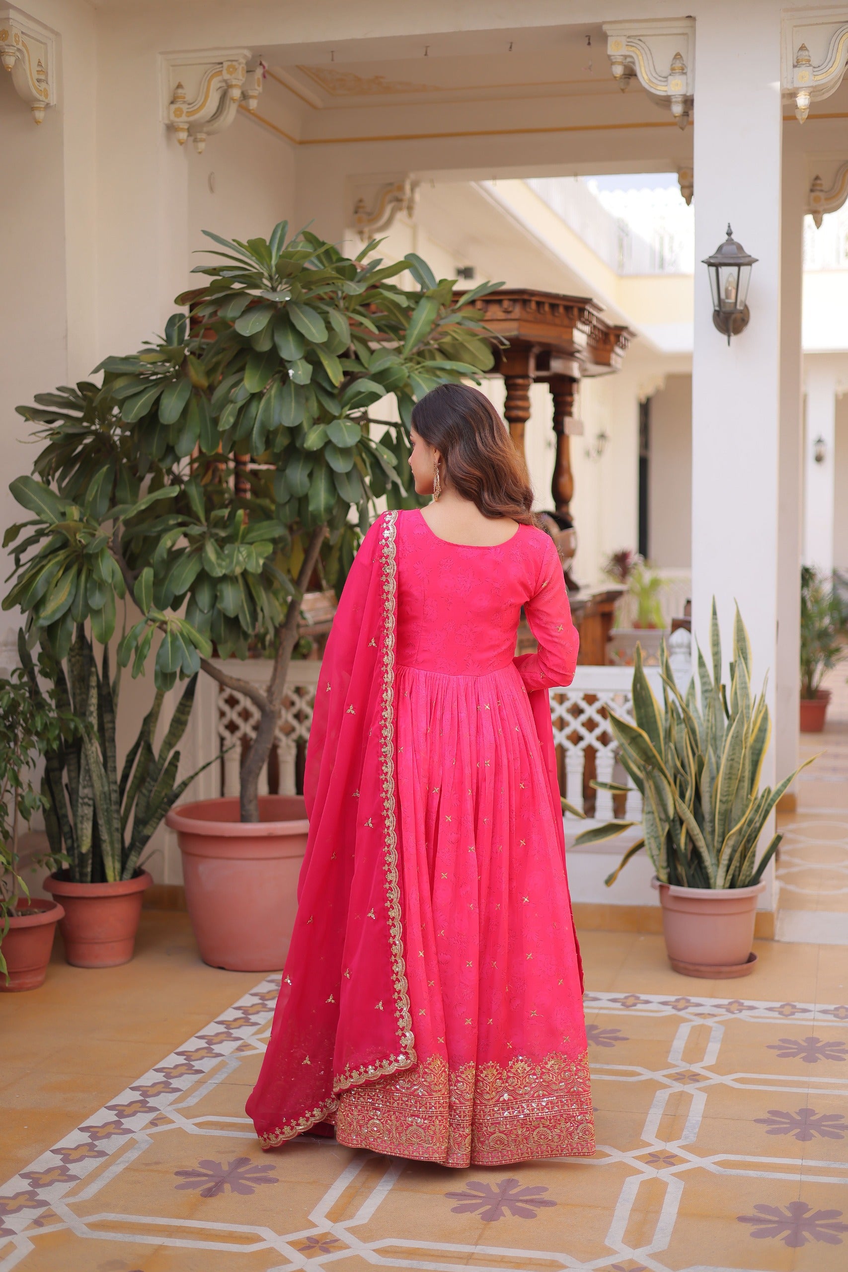 Woman in a pink traditional outfit standing in a decorated indoor setting with plants and architectural elements.