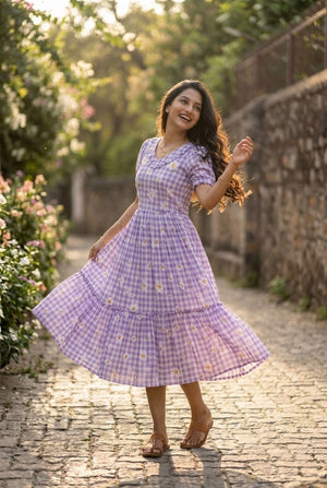 Woman in a lavender checkered dress standing on a stone path with greenery around.