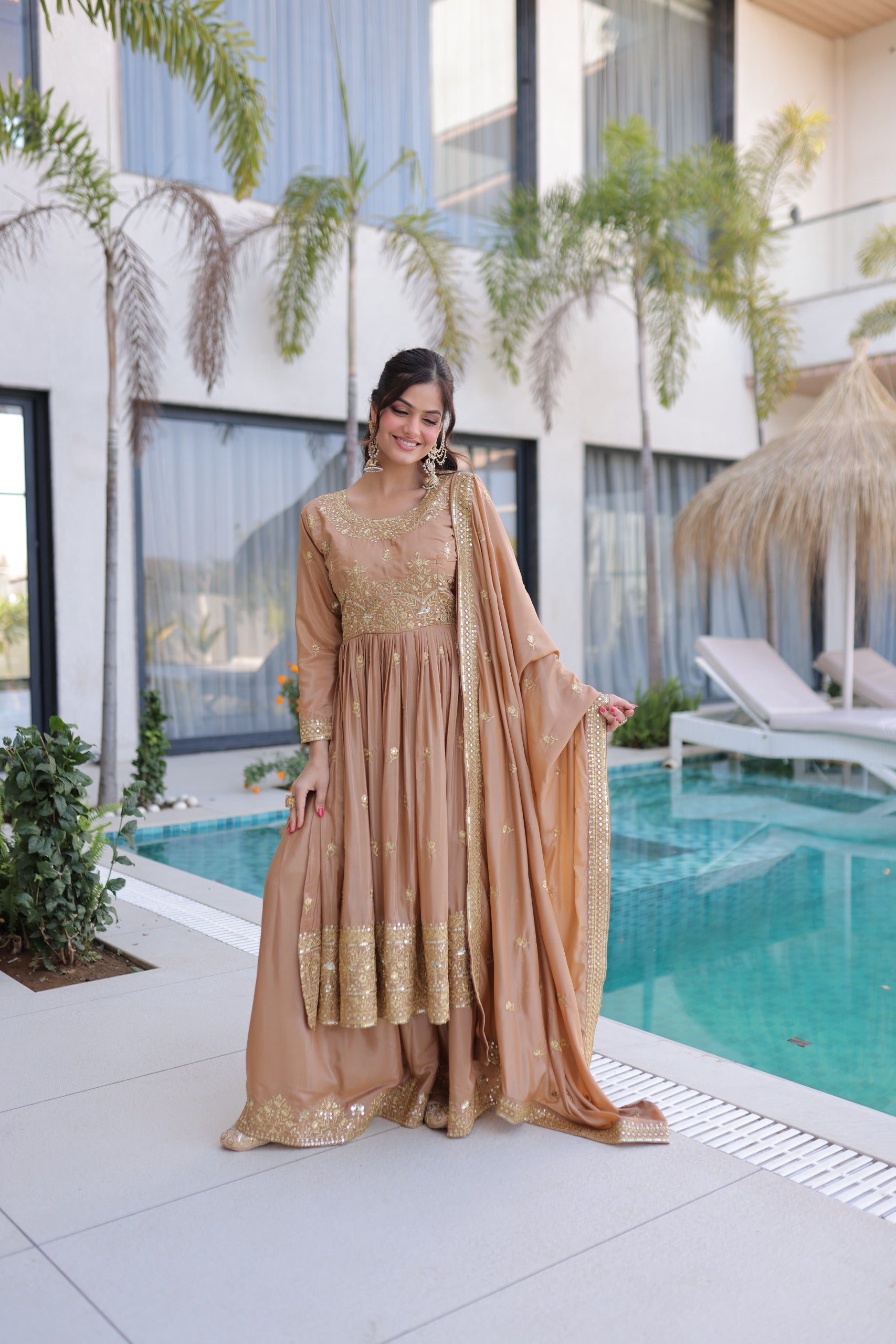 Woman in a beige and gold traditional outfit standing by a pool with modern building and plants in the background