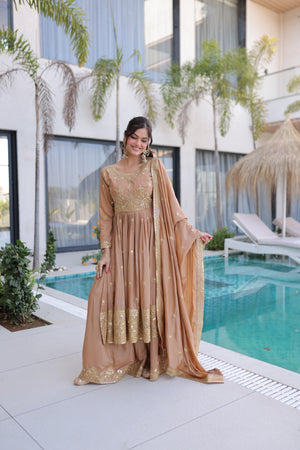 Woman in a beige and gold traditional outfit standing by a pool with modern building and plants in the background