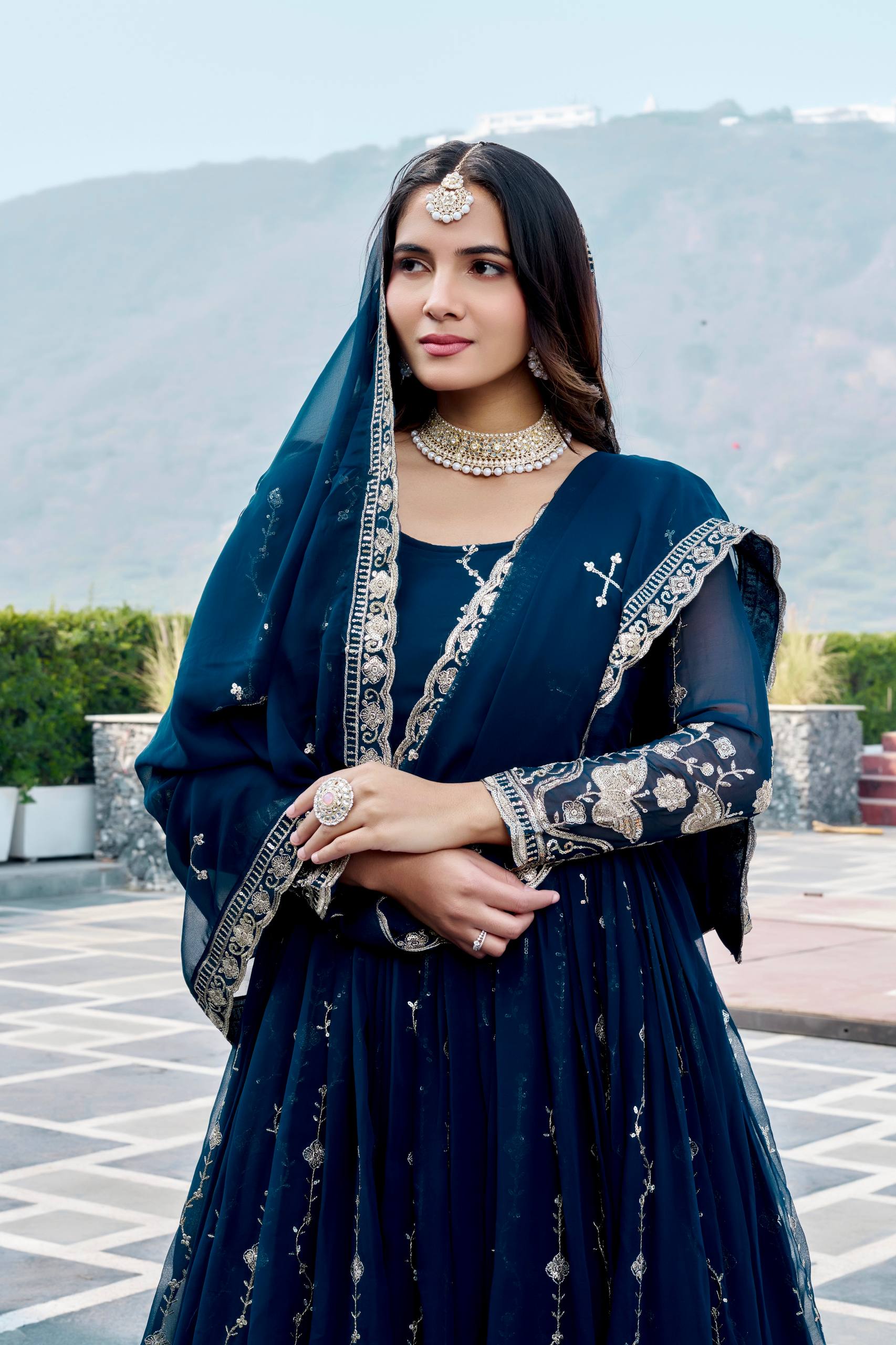 Woman in a dark blue traditional outfit with embroidery, standing outdoors with mountains in the background.