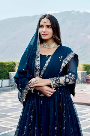 Woman in a dark blue traditional outfit with embroidery, standing outdoors with mountains in the background.