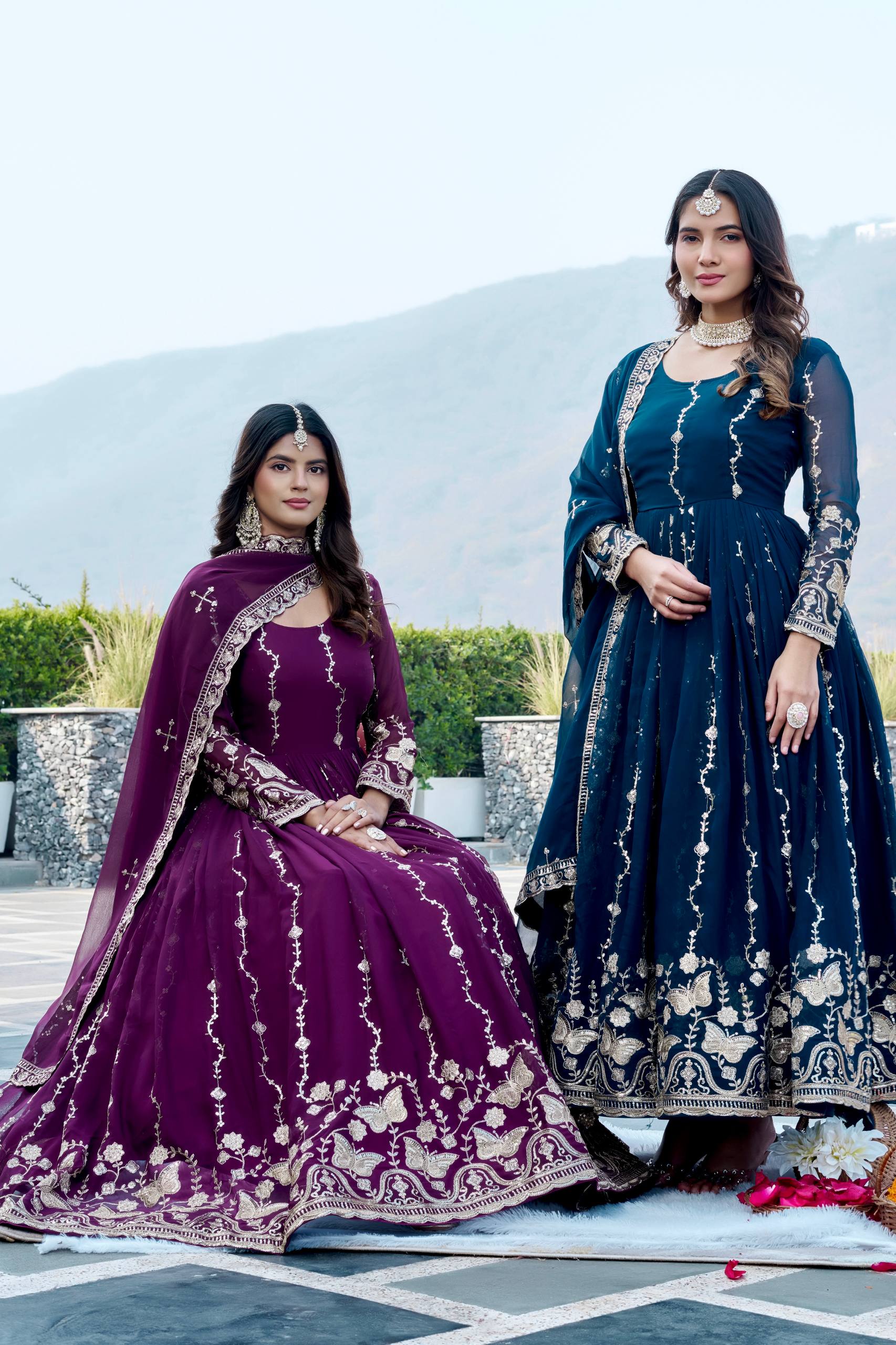 Two women in traditional outfits standing outdoors with a scenic background.