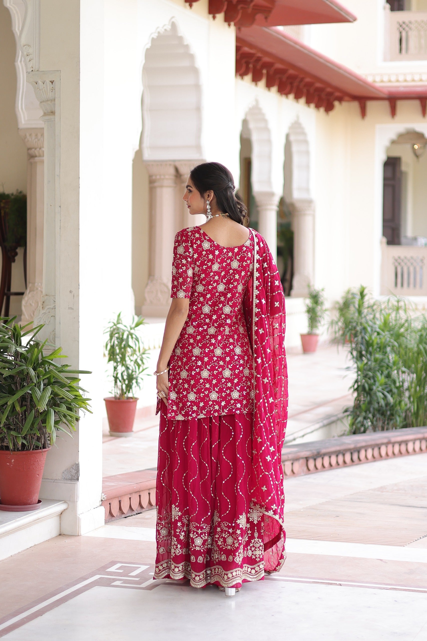 Woman in a red traditional outfit standing in a decorative outdoor setting with plants and architectural elements.