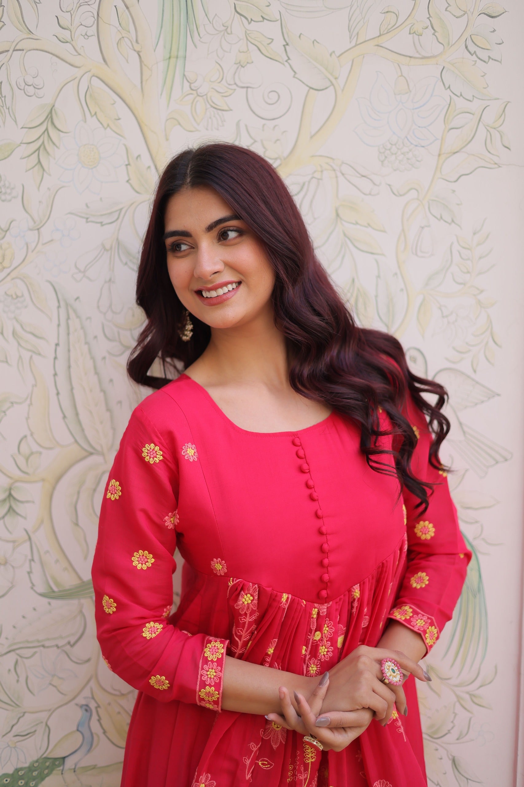 Woman wearing a red dress with gold patterns against a decorative wall.