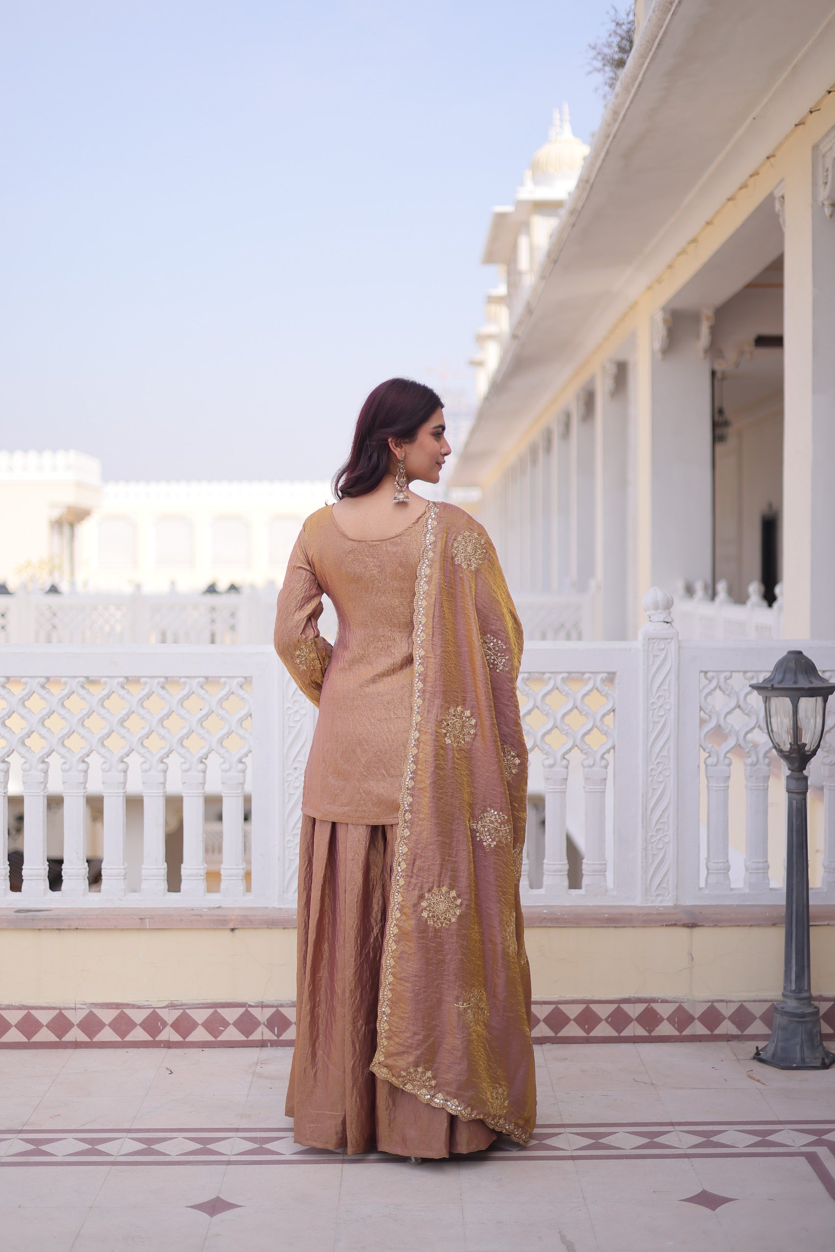 Woman in a traditional outfit standing on a balcony with white columns and a clear sky.