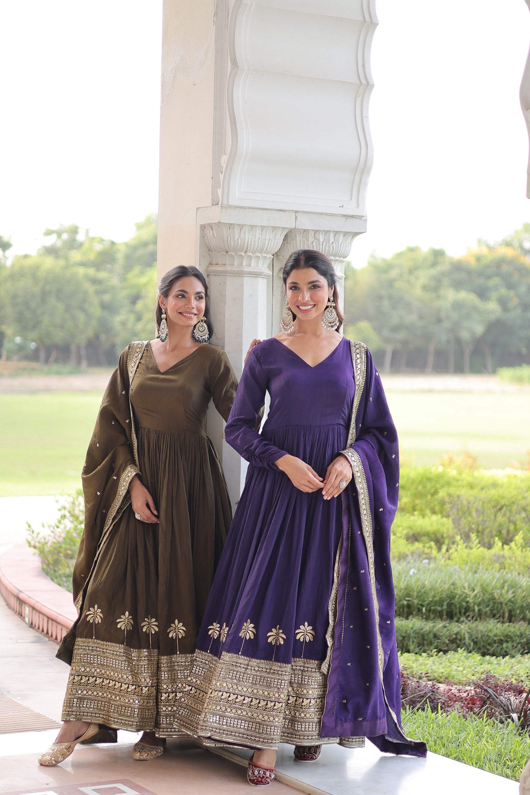 Two women in traditional outfits standing outdoors with greenery in the background