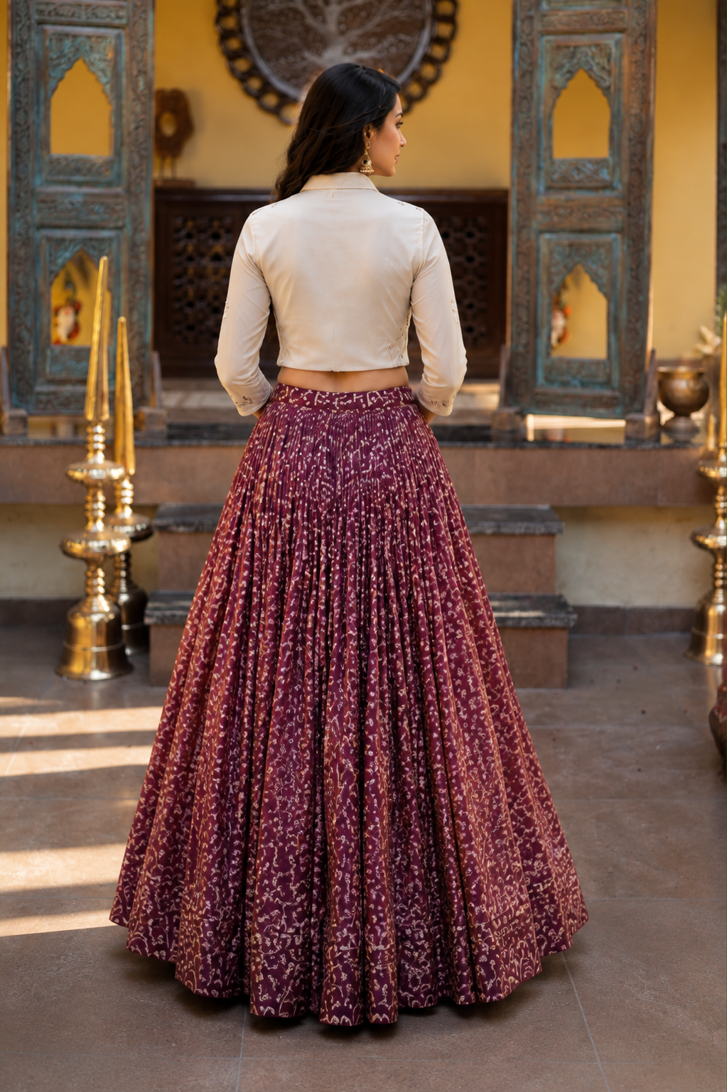A woman viewed from behind wearing a cream long-sleeve crop top and a voluminous maroon skirt with a dense gold floral pattern, standing in a traditional courtyard with ornate wooden doors.