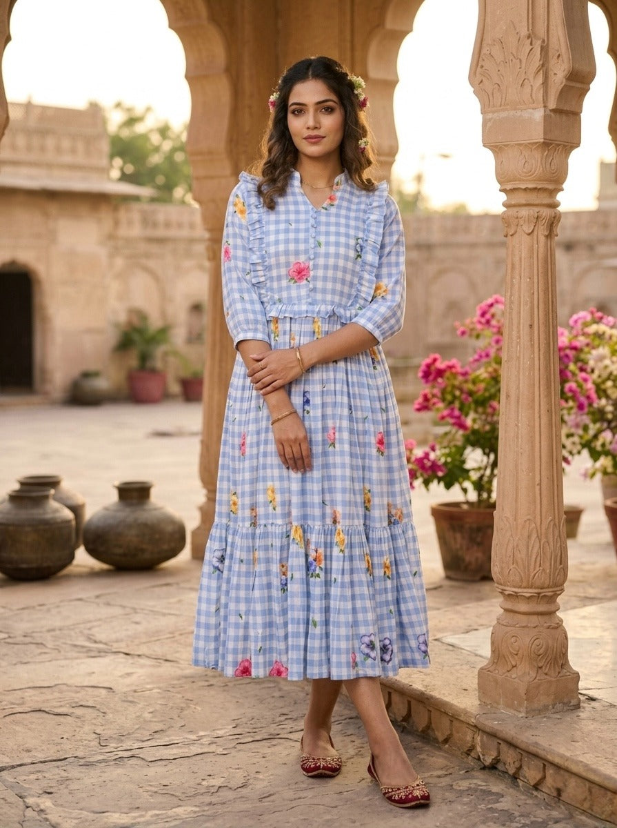 Woman in a blue checkered dress with floral embroidery standing in an outdoor setting with architectural elements and flowers.