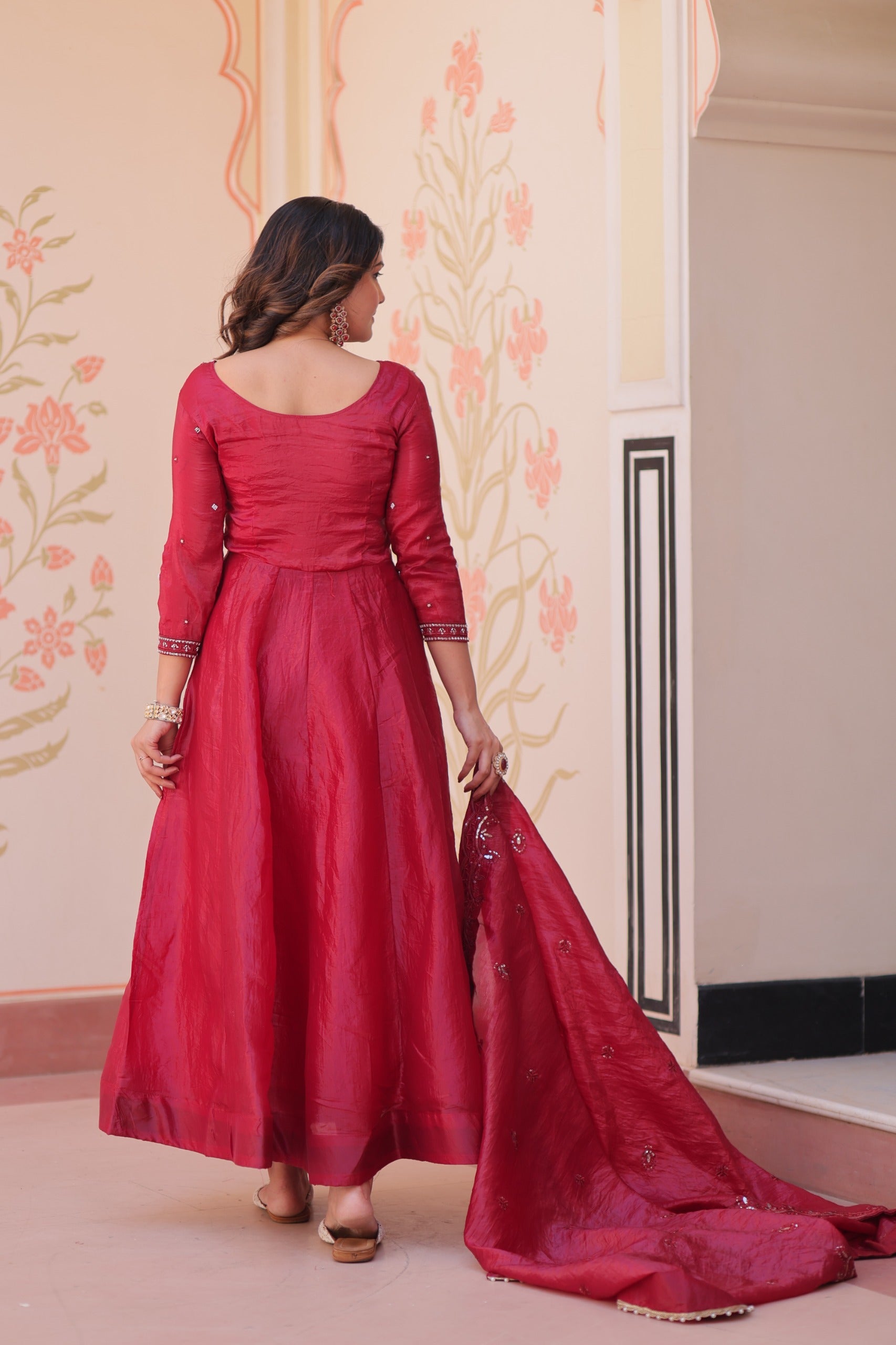 Woman in a red anarkali suit standing in a room with floral wallpaper.