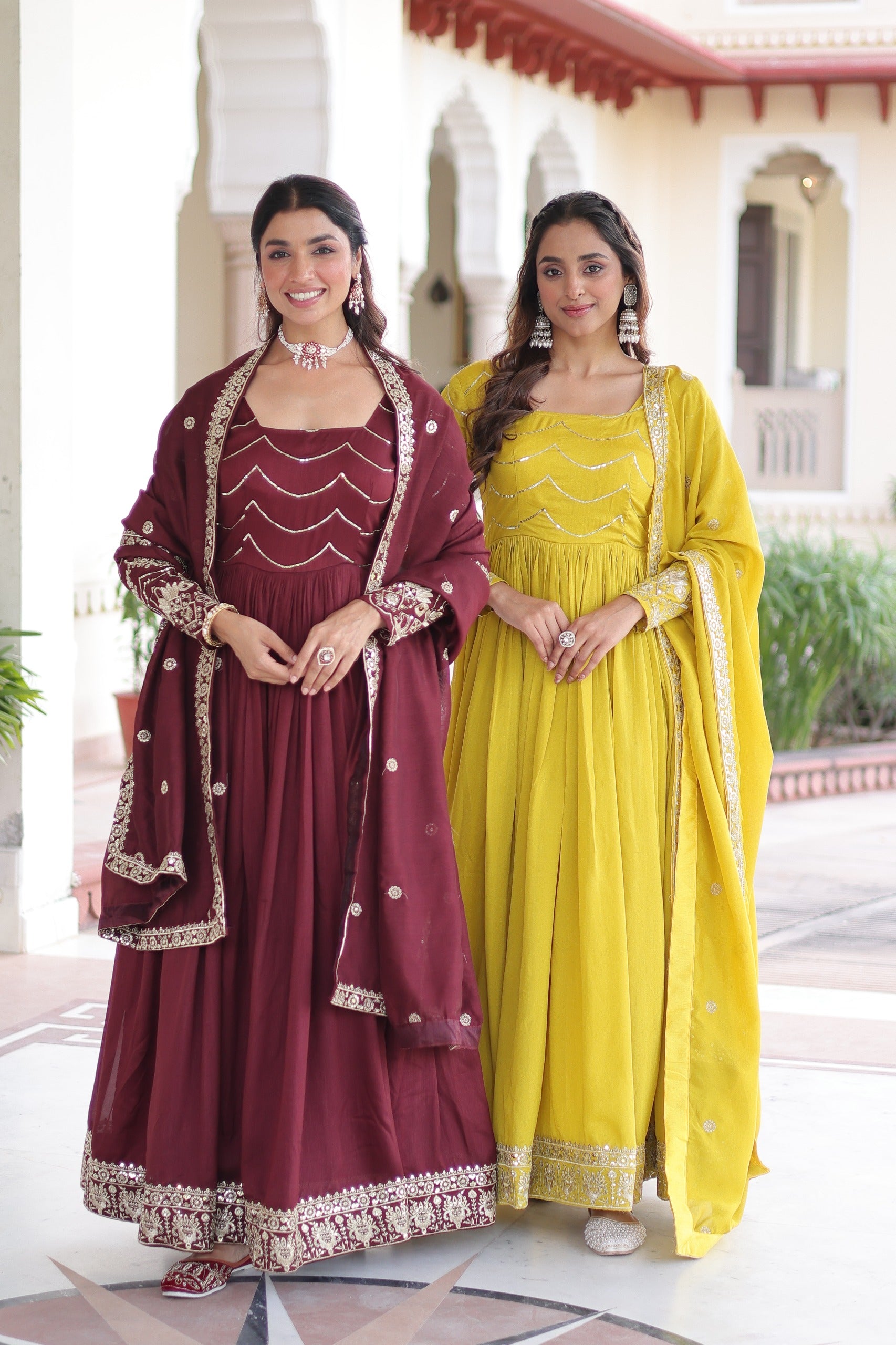Two women in traditional maroon and yellow dresses standing in front of a building.
