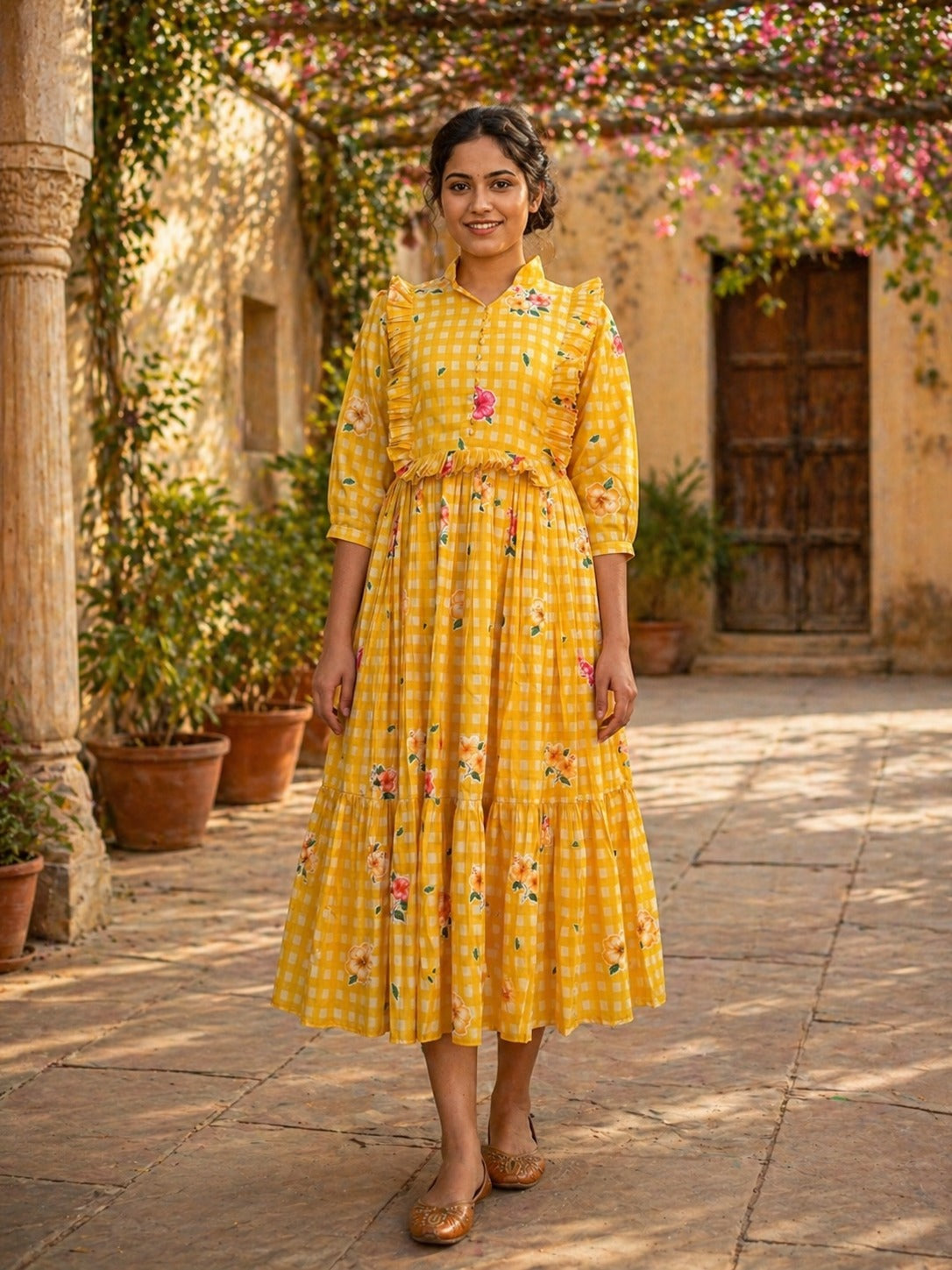 Woman in a yellow checkered dress standing in an outdoor setting with plants and a building in the background.