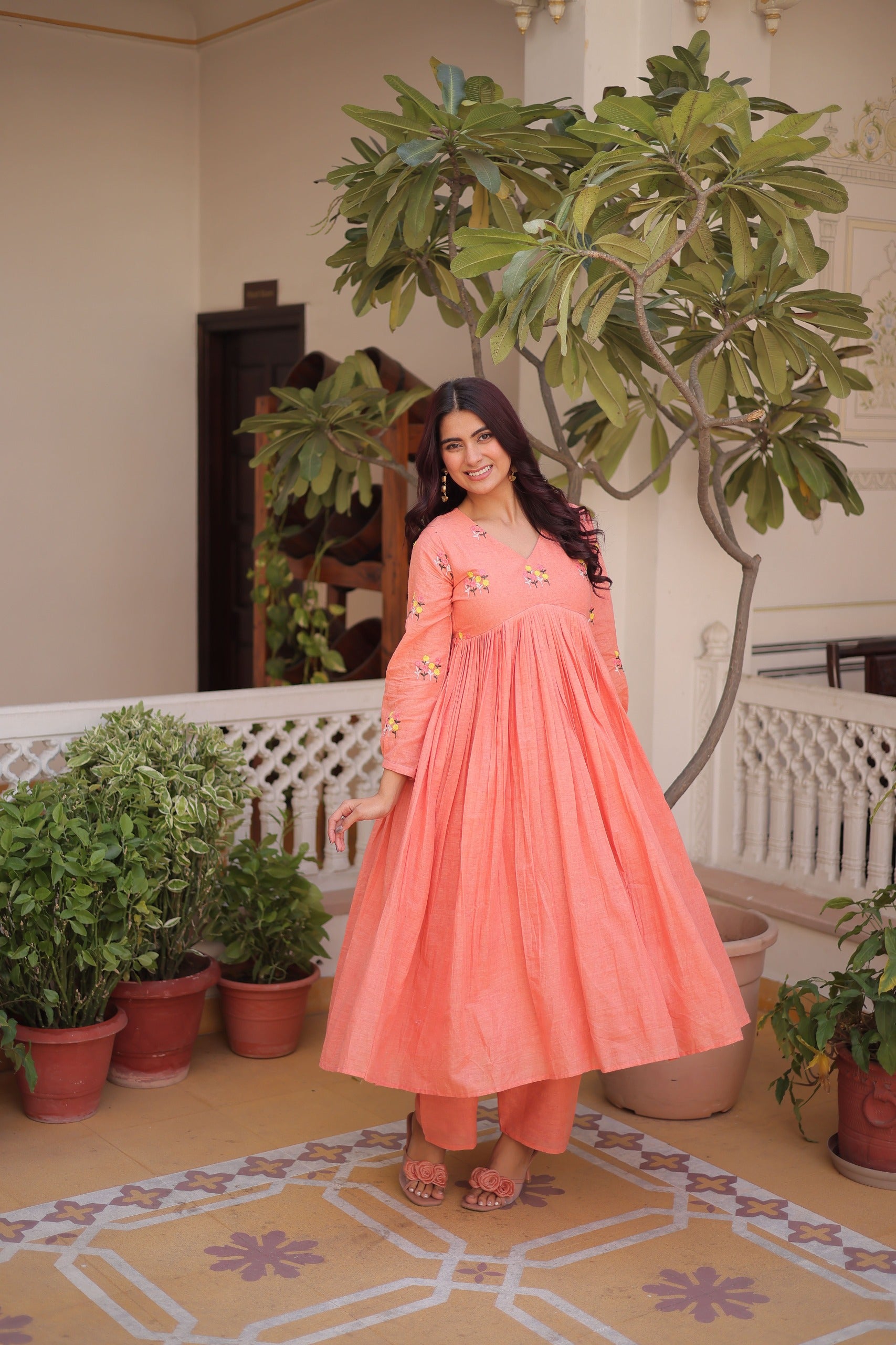 Woman in a peach-colored dress standing in a room with plants and decorative tiles.