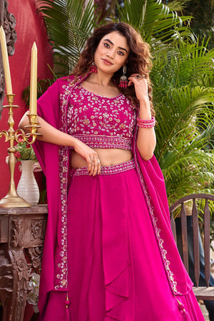 Woman in a pink traditional outfit with floral patterns, standing outdoors with greenery and a decorative table in the background.