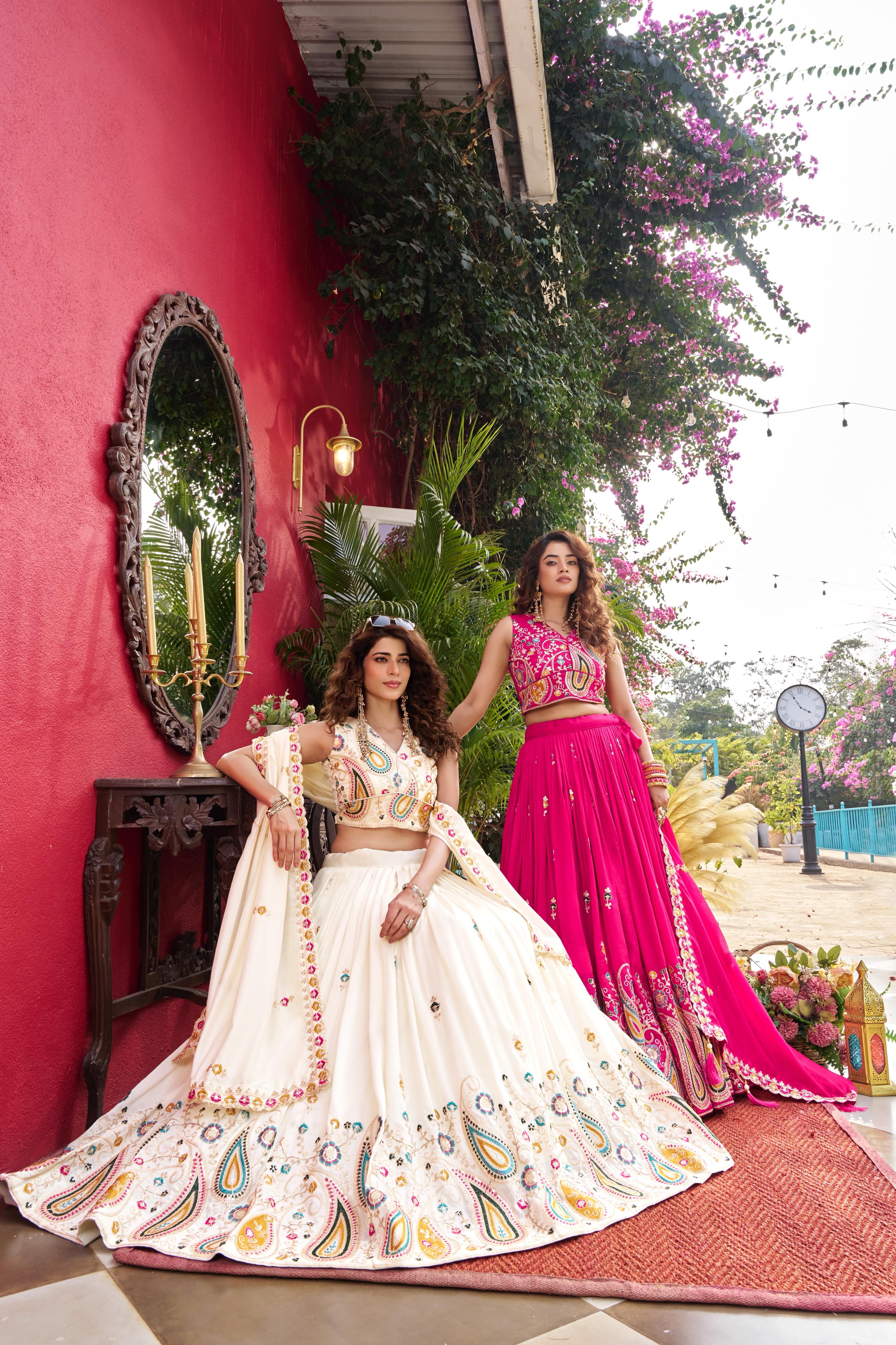 Two women in traditional outfits standing in front of a red wall with plants.