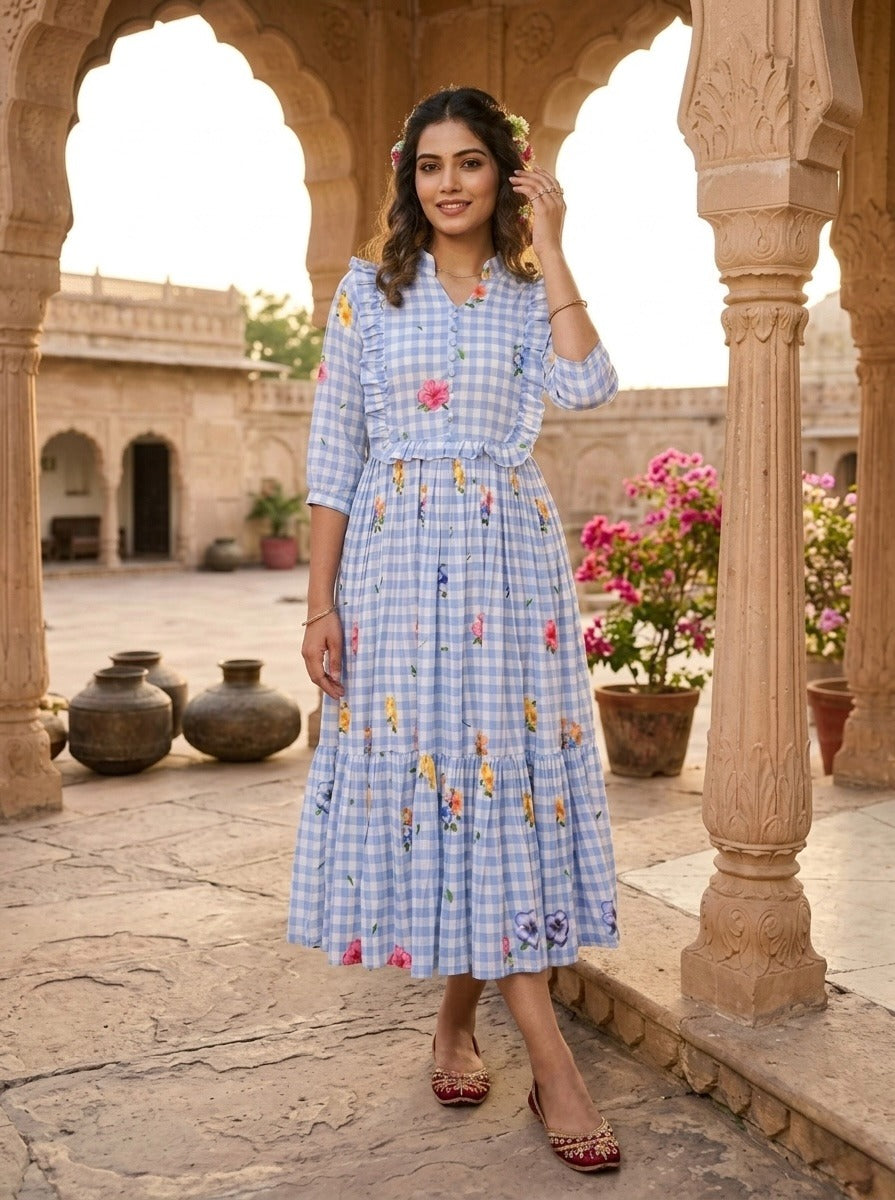 Woman in a blue checkered dress with floral embroidery standing in an outdoor setting with arches and plants.