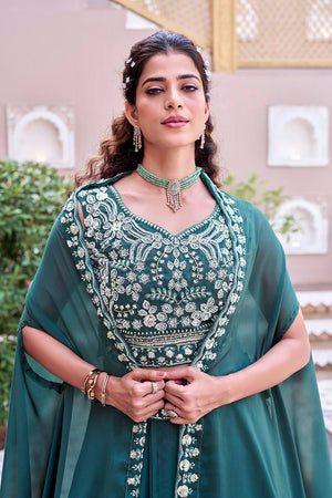 Woman wearing a green embroidered traditional outfit with jewelry against a decorative wall.