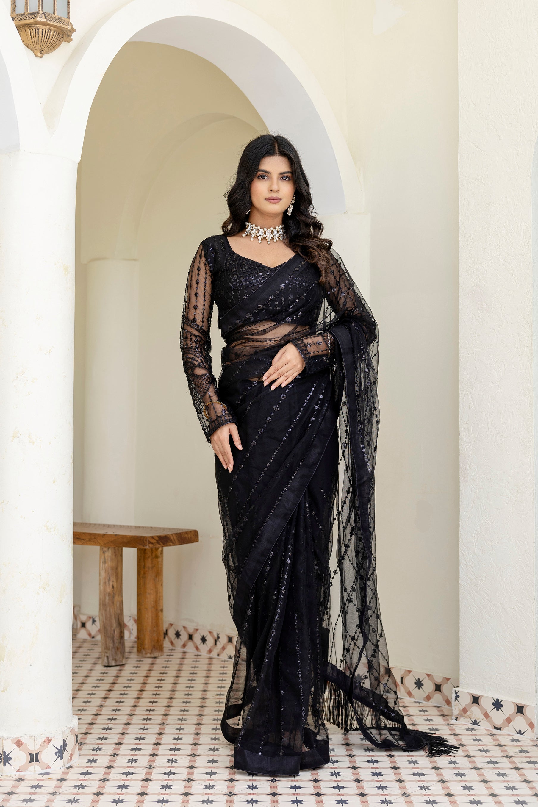Woman in a black saree standing in a well-lit room with white walls and a wooden table.