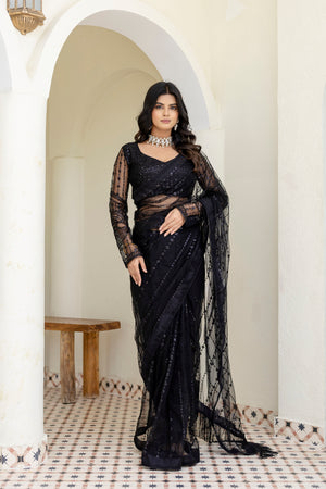 Woman in a black saree standing in a well-lit room with white walls and a wooden table.