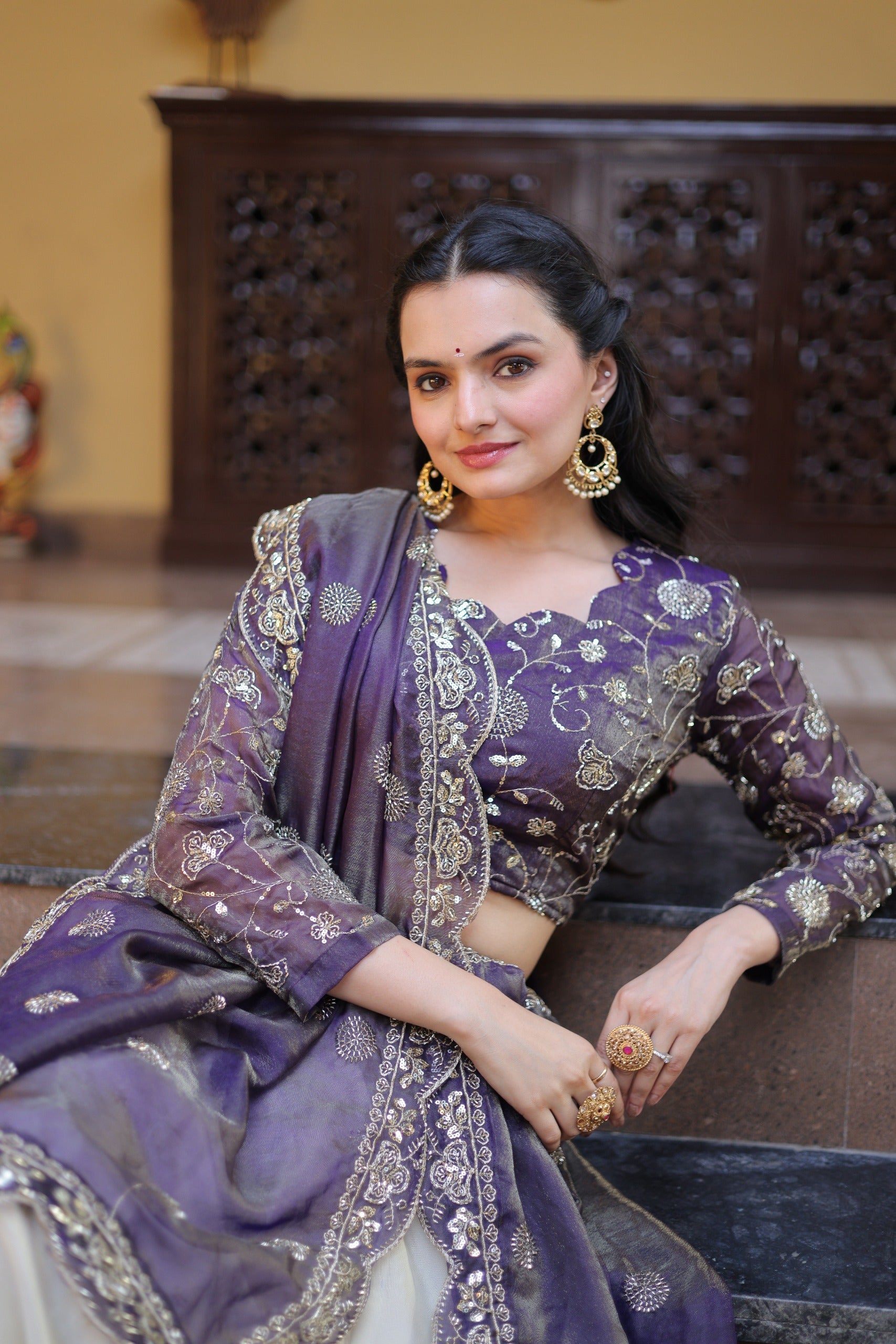 Woman in a traditional purple and gold outfit sitting on a decorative chair.